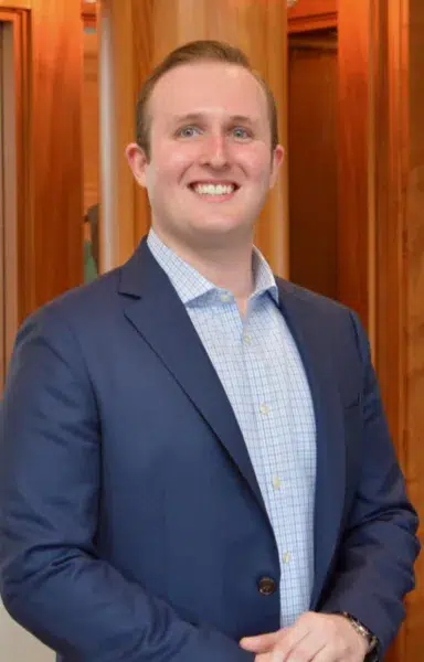 A man in a blue suit and checked shirt smiles while standing indoors in front of wooden paneling.