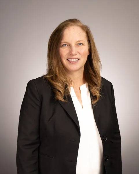 A woman with long, light brown hair in a black blazer and white blouse smiles at the camera against a neutral gray background.