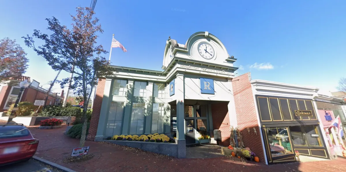 A brick building with a large clock above the entrance, an American flag on a pole, yellow flowers, and blue sky in the background.