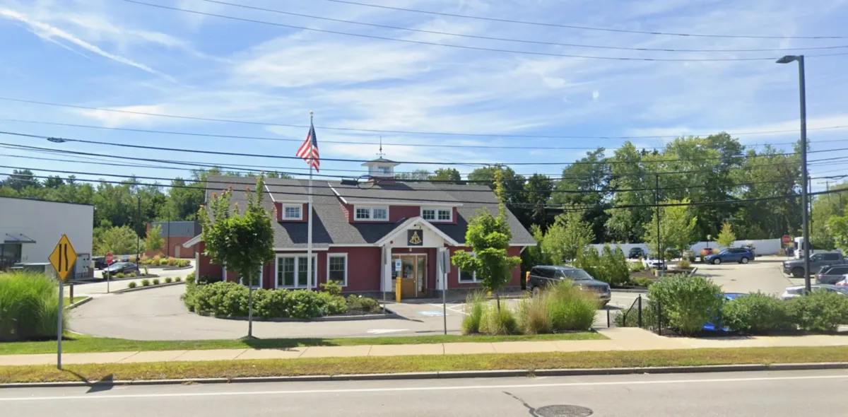 A red and white building with an American flag on top, surrounded by greenery and parked cars, located along a street under a blue sky.