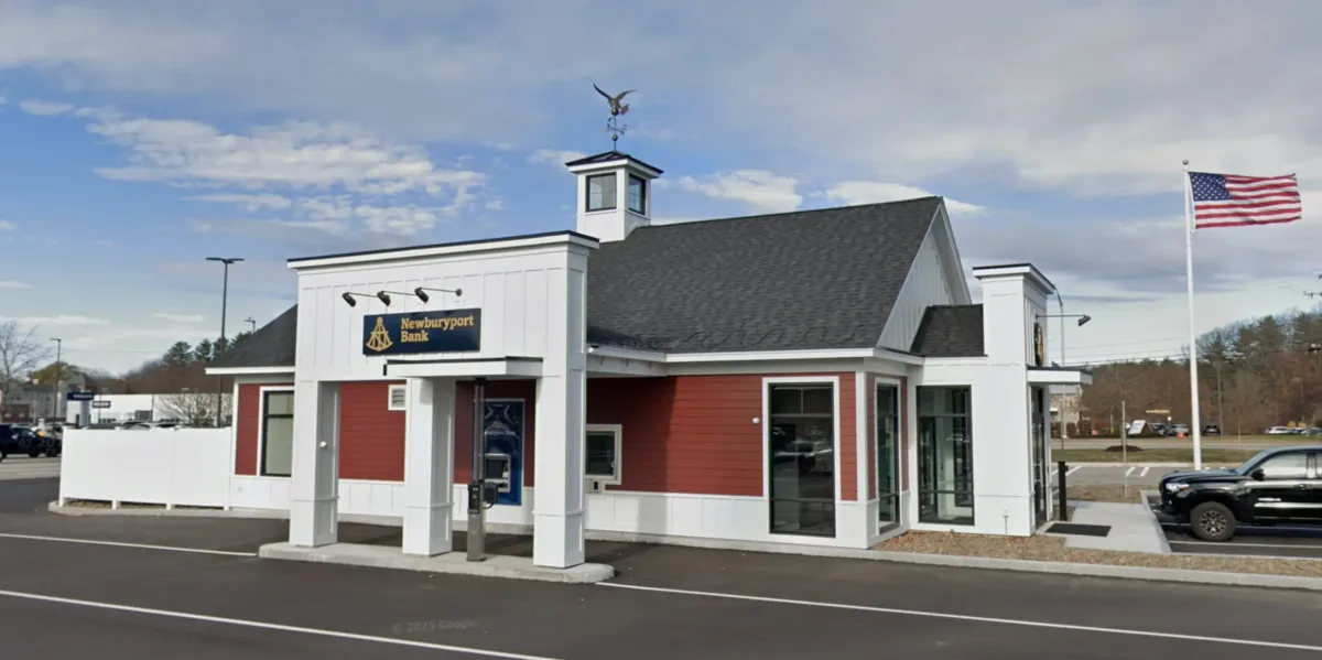 A small red and white bank building with a drive-thru, an American flag, and an eagle statue on the roof under a partly cloudy sky.