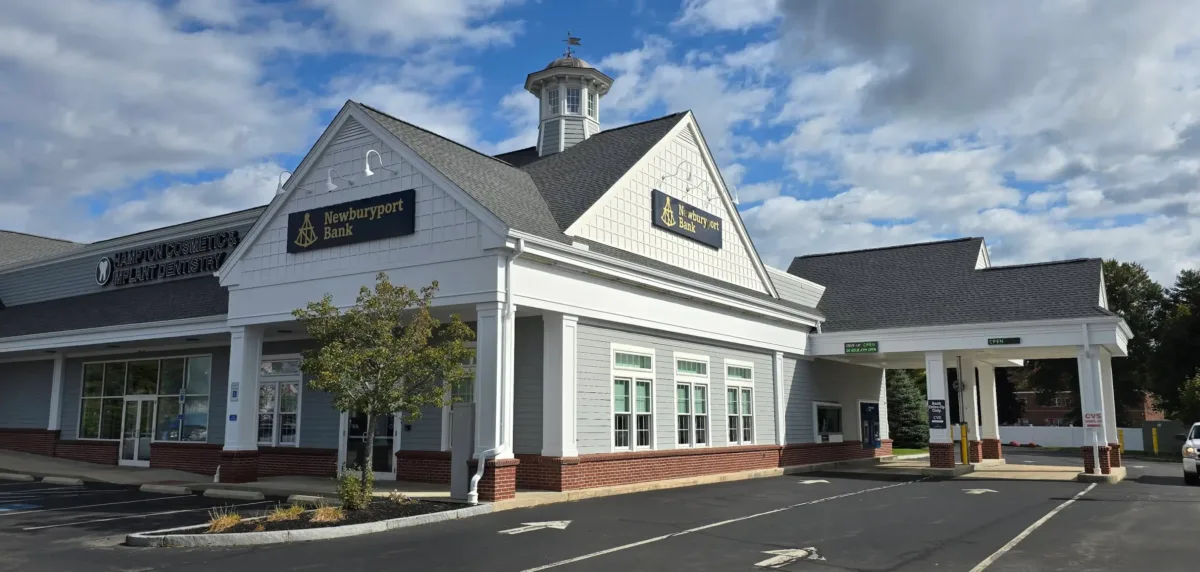 A white bank building with a drive-thru, large windows, and a cupola on the roof, set against a blue sky with scattered clouds.
