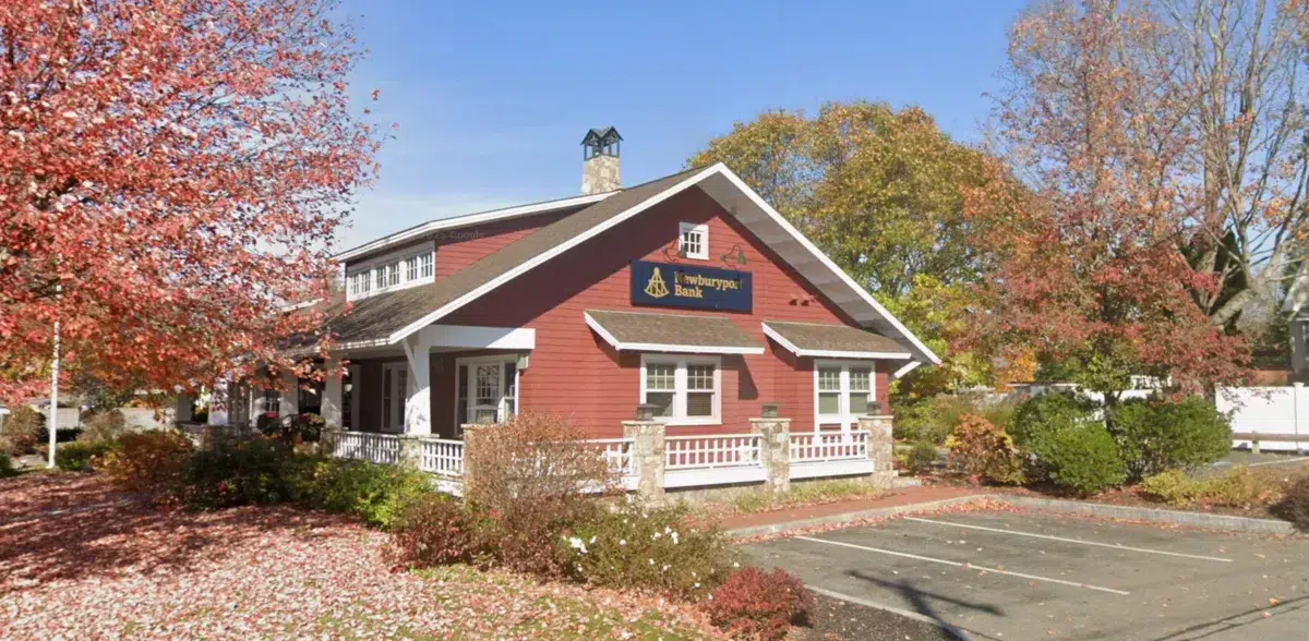 A red building with white trim and a porch, surrounded by autumn trees and a nearly empty parking lot under a clear blue sky.
