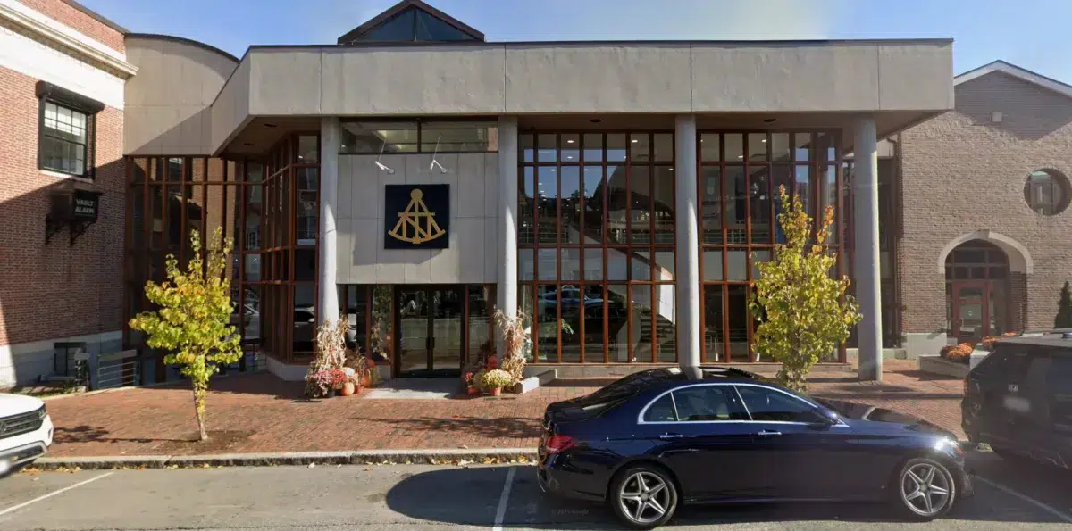 Modern two-story building with large glass windows, a nautical symbol above the entrance, fall decorations, and a parked black car in front on a brick-paved sidewalk.