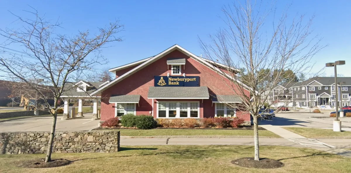 A red building with a sign reading Newburyport Bank, surrounded by trees and shrubs under a clear blue sky.