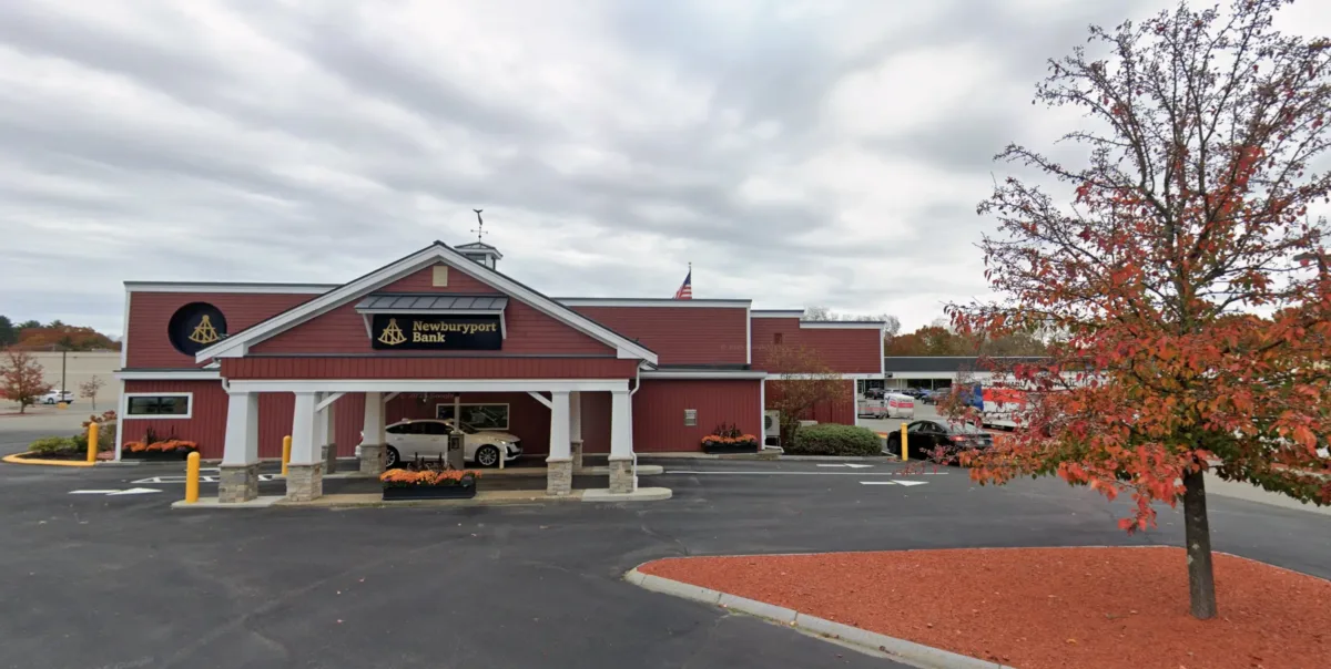 A red building with a white canopy and Newburyport Bank sign, surrounded by a parking lot and autumn trees under a cloudy sky.