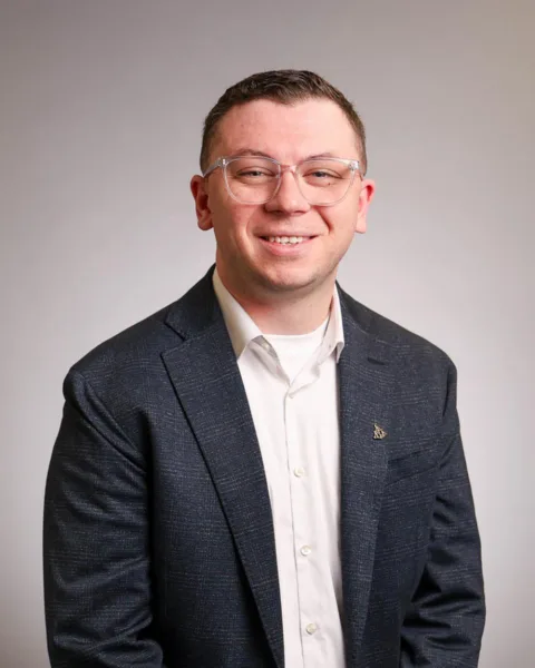 A man wearing glasses, a white shirt, and a dark blazer smiles at the camera against a plain light background.