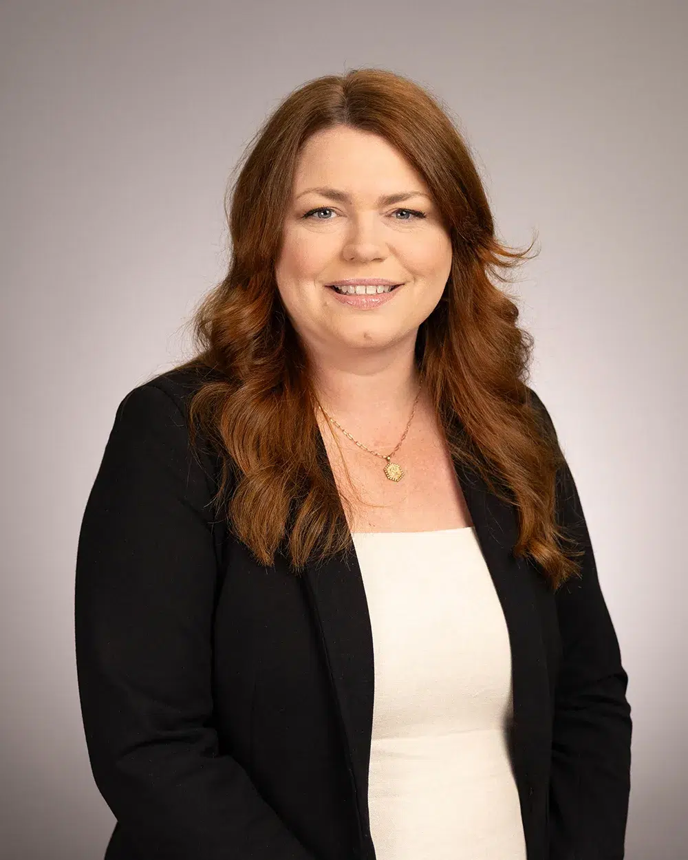 A woman with long, wavy brown hair wearing a black blazer over a white top, smiling at the camera against a plain light gray background.