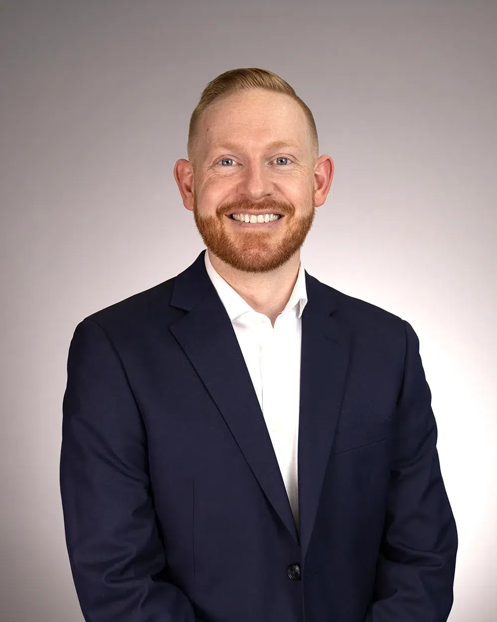 A smiling man with short red hair and a beard wears a navy suit jacket over a white shirt, standing against a plain light gray background.