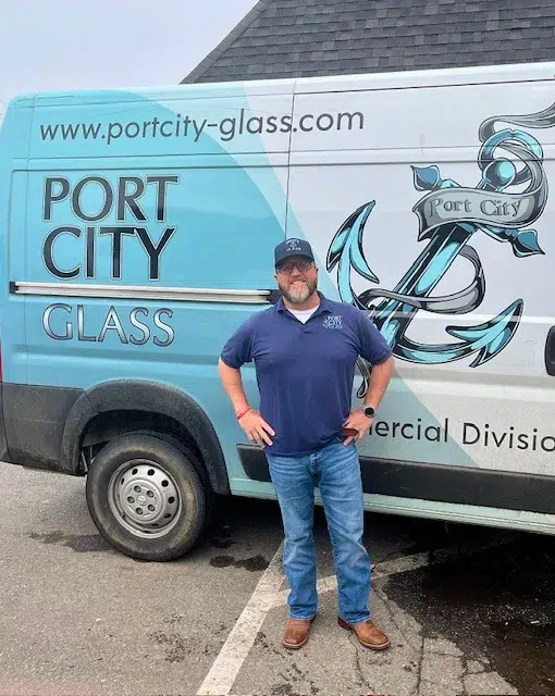 A man in a navy polo and cap stands in front of a blue Port City Glass van with an anchor logo and website text.
