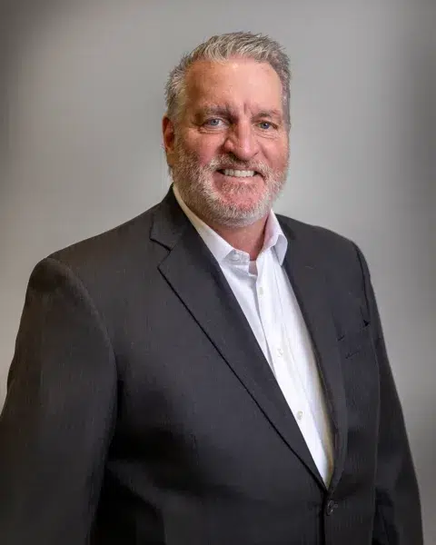 A middle-aged man with gray hair and a beard, wearing a dark suit and white shirt, smiles at the camera against a plain gray background.