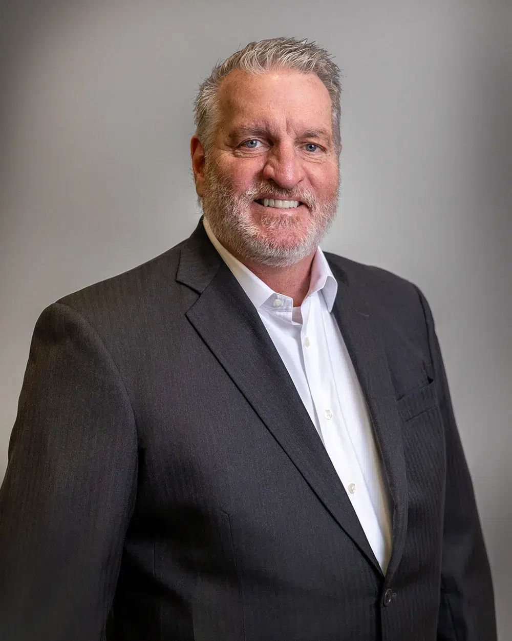 A middle-aged man with gray hair and a beard, wearing a dark suit and white shirt, smiles at the camera against a plain gray background.