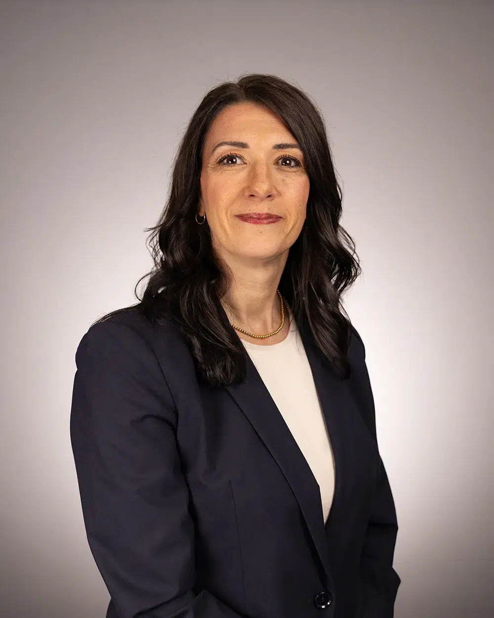 A woman with long dark hair, wearing a navy blazer over a light top, stands against a plain, light gray background, smiling slightly at the camera.