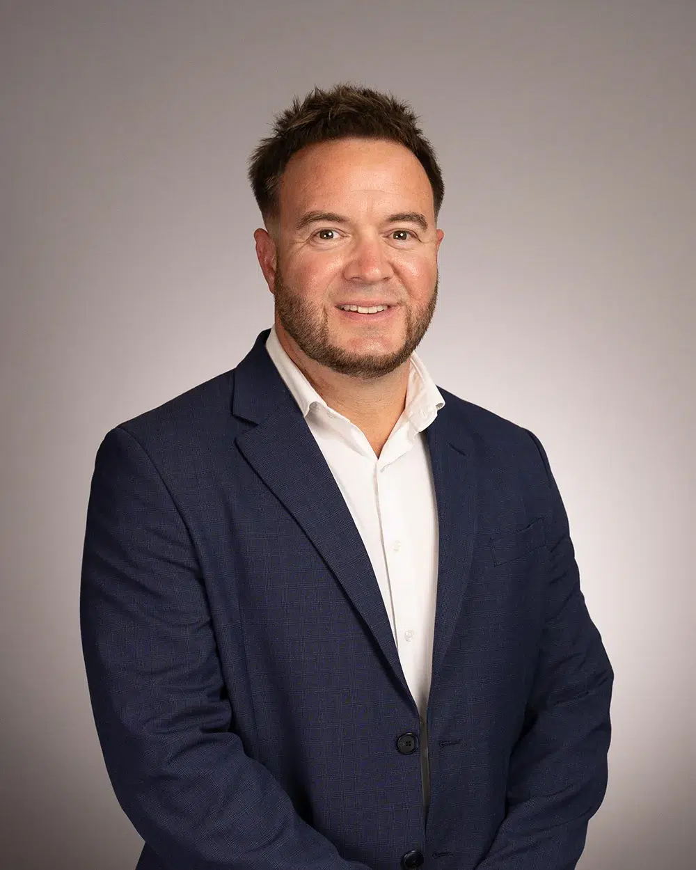 A man with short brown hair and a trimmed beard, wearing a navy blue suit and white shirt, stands smiling against a plain light gray background.