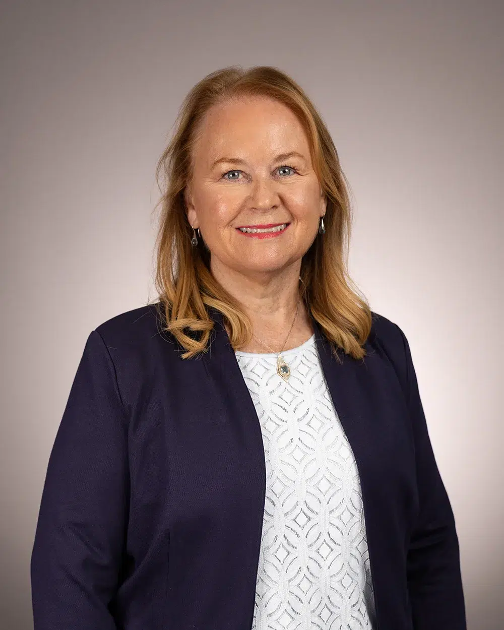 A woman with shoulder-length blonde hair, wearing a navy blazer over a white patterned shirt, smiles at the camera against a plain, light-colored background.