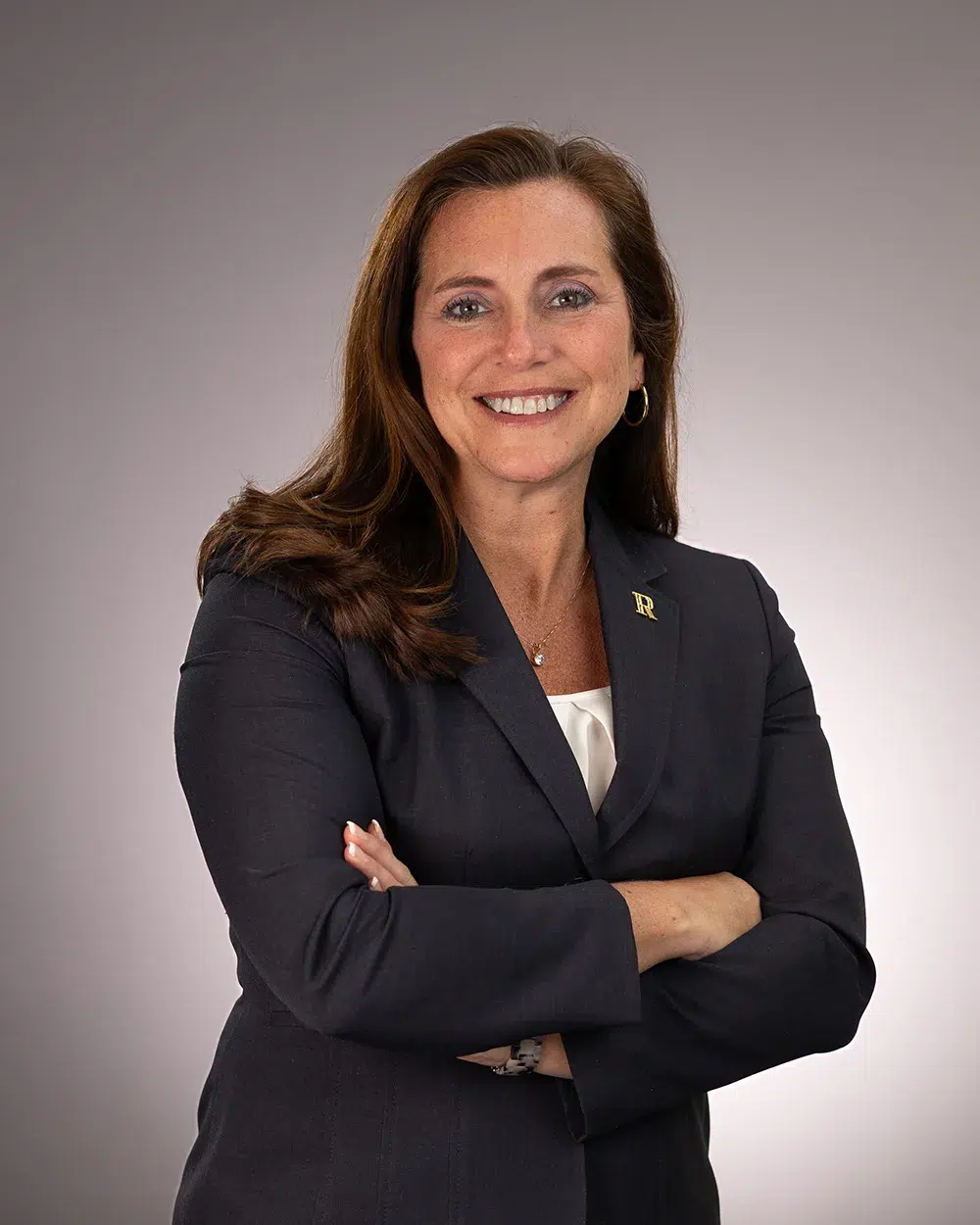 A woman with long brown hair in a dark blazer smiles with arms crossed against a plain gray background.
