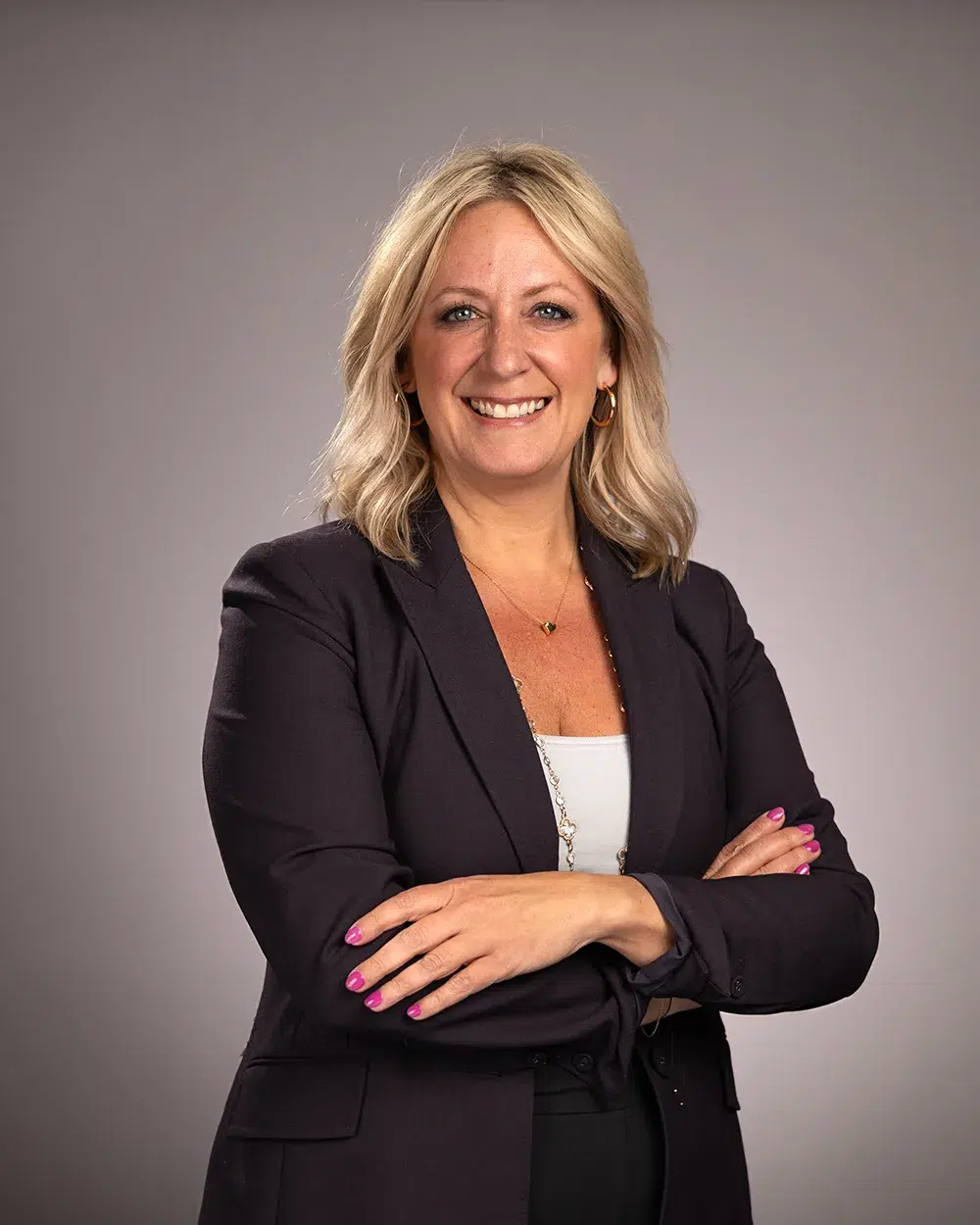 A smiling woman with blonde hair in a dark blazer stands with arms crossed against a plain, light gray background.