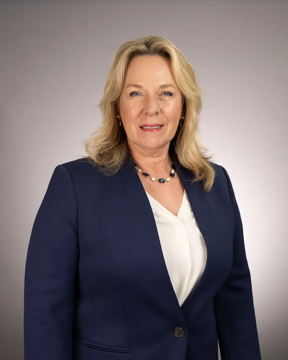 A woman with blonde hair, wearing a navy blazer over a white blouse and a beaded necklace, stands against a plain light gray background, smiling slightly.