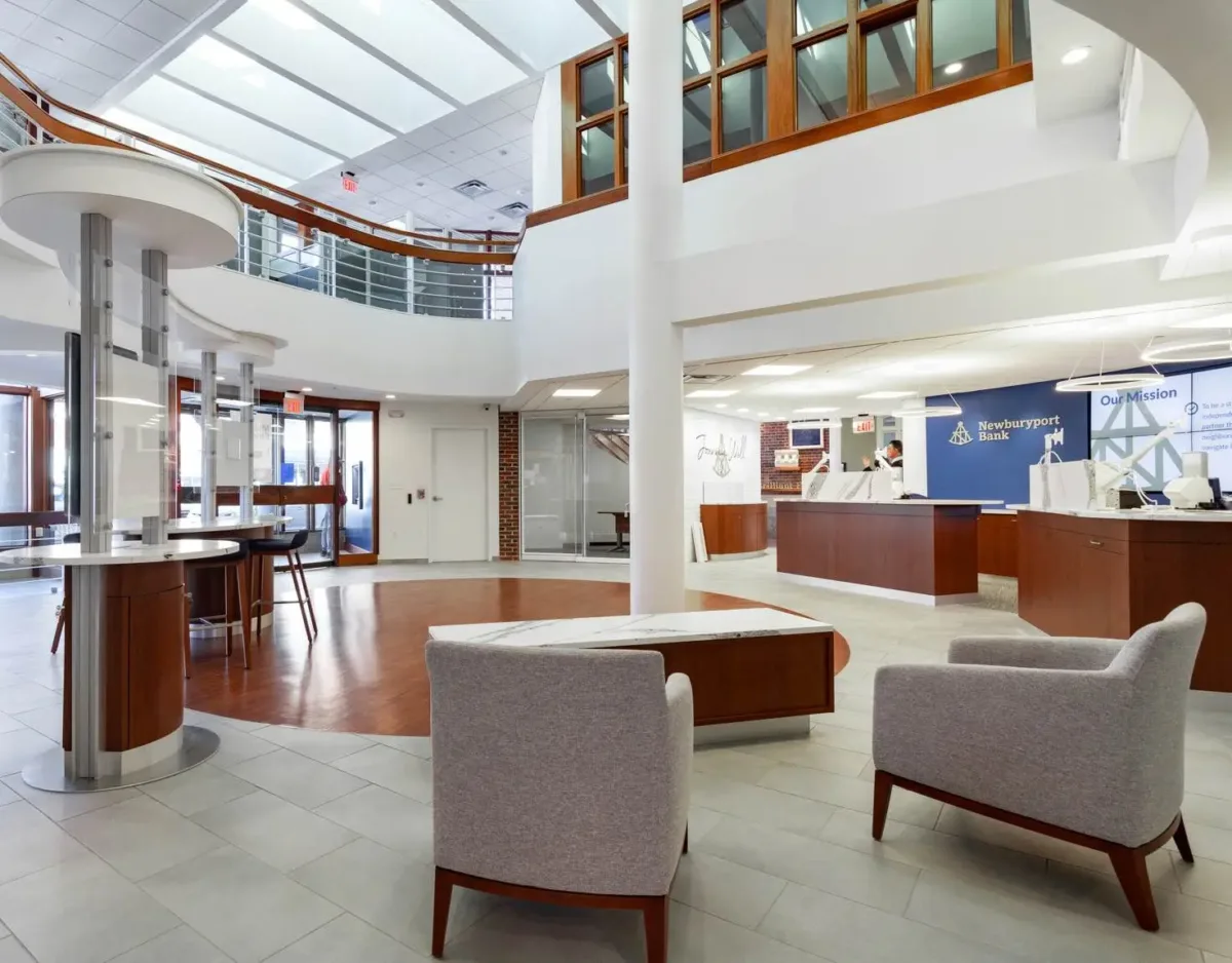 Modern bank interior with large windows, a curved staircase, information desks, seating area with gray chairs, and bright natural lighting.