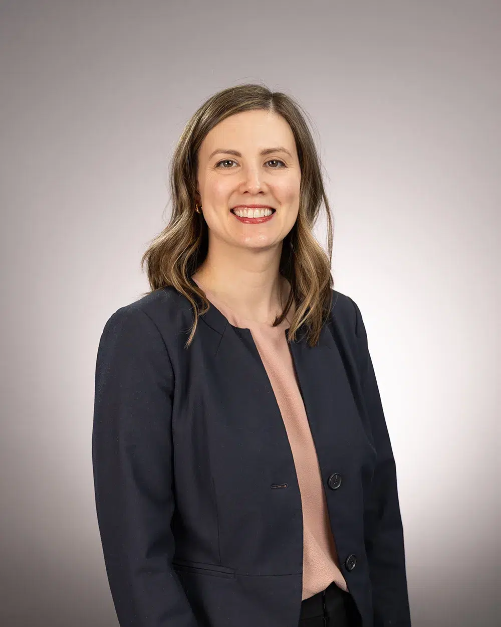 A woman with shoulder-length brown hair, wearing a navy blazer and light pink top, smiles in front of a plain gray background.
