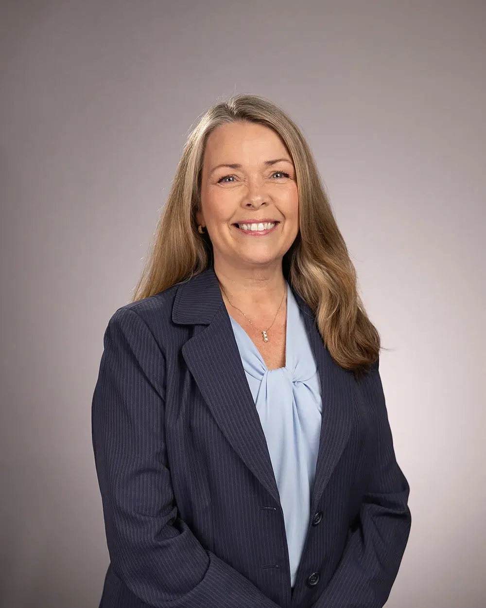 A woman with long blonde hair wearing a navy blazer and light blue blouse smiles in front of a plain gray background.