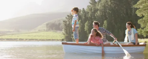A family of four enjoys a sunny day on a rowboat in a lake, surrounded by trees and mountains. One child stands looking ahead, while the others sit and smile.