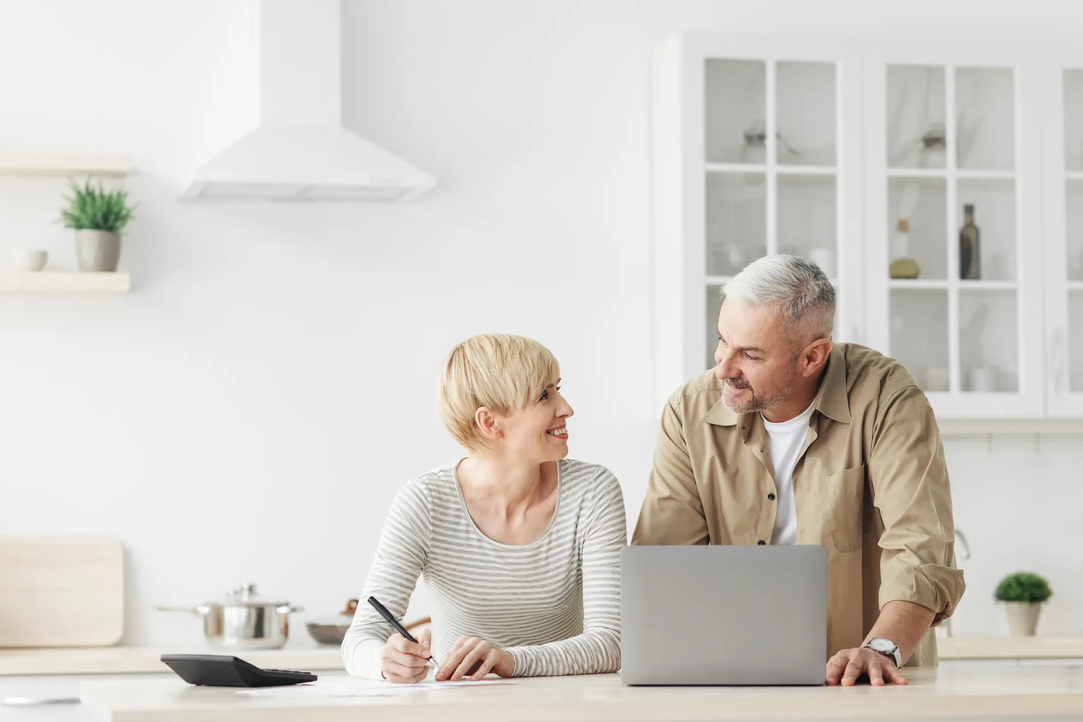 A woman writes in a notebook while smiling at a man standing beside her. They are in a bright kitchen with a laptop and tablet on the counter.