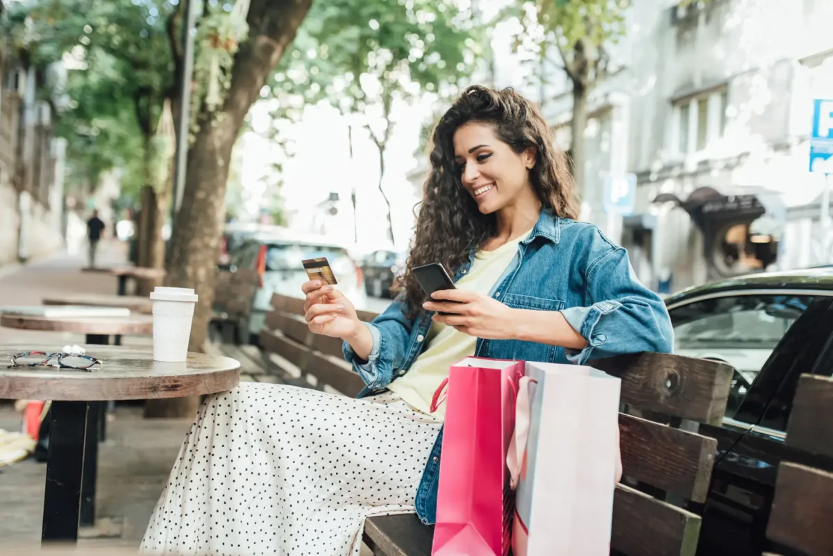 A woman sits on a bench outdoors, smiling while holding a credit card and smartphone. Shopping bags and a coffee cup are on the table beside her.