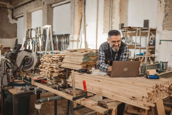 A man in a workshop smiles while working on a laptop, surrounded by stacks of wood, tools, and carpentry equipment.