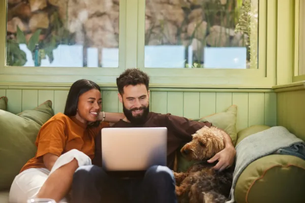 A smiling couple sits on a cozy couch with a laptop, while their brown dog rests beside them in a sunlit room.