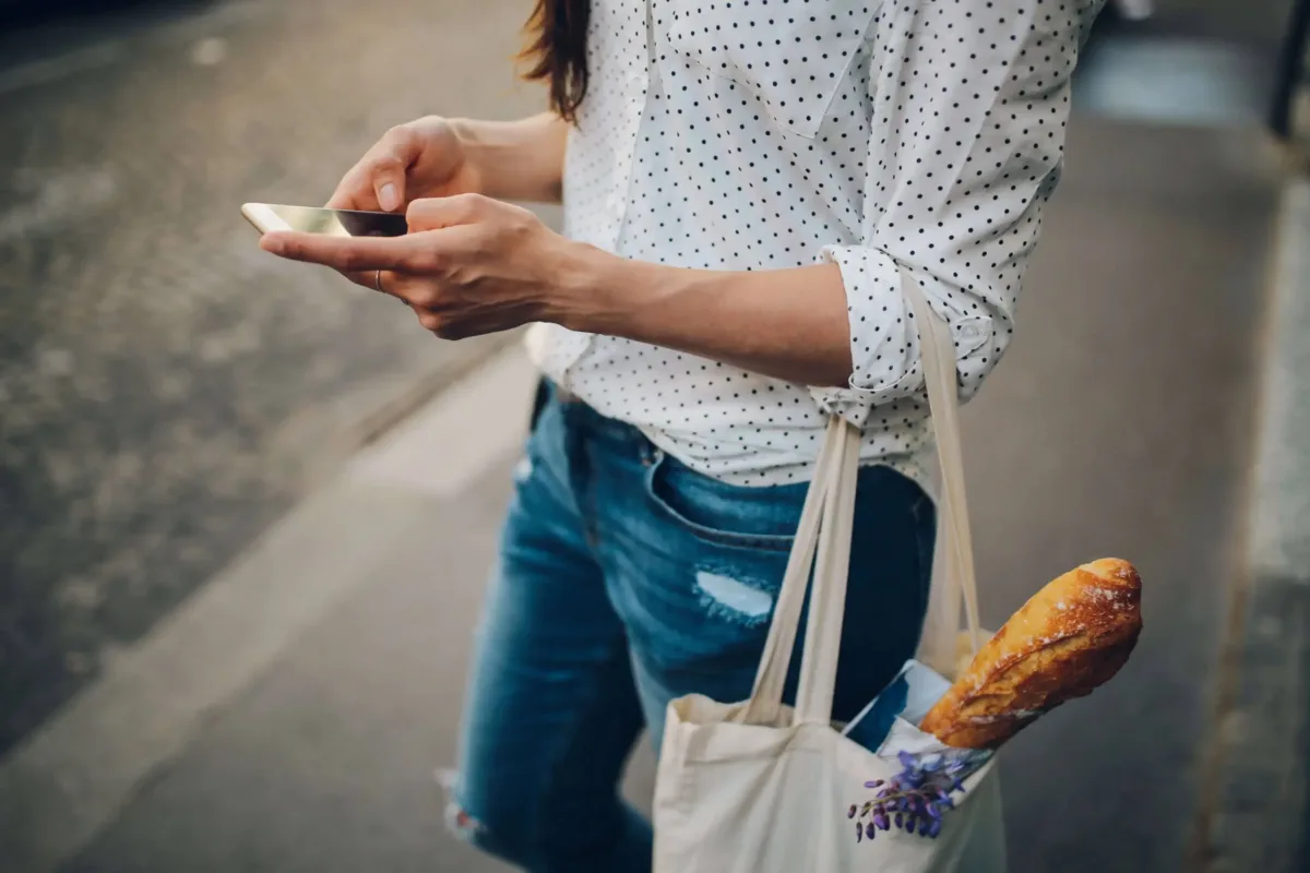 A person in a white polka dot shirt and jeans walks on a sidewalk, using a smartphone, with a baguette in a tote bag hanging from their shoulder.