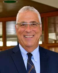 Smiling older man with short gray hair, glasses, and a dark suit with a striped tie, standing indoors in front of wooden framed windows.