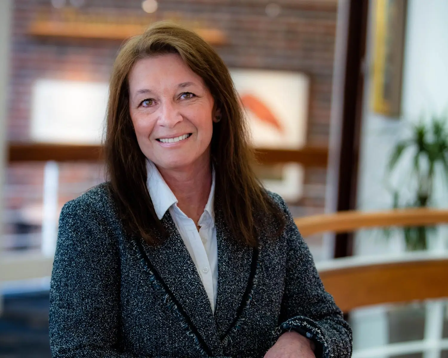 A woman with long brown hair wearing a gray blazer and white shirt smiles while standing indoors in a brightly lit modern space.