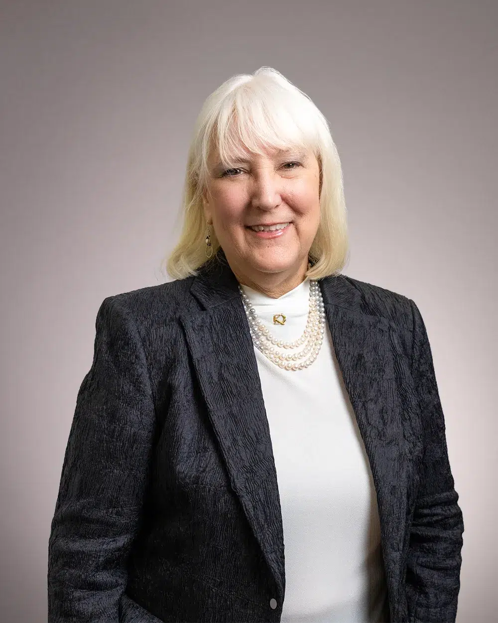 A smiling older woman with white hair wears a dark textured blazer, white top, and layered pearl necklaces, standing against a plain light background.