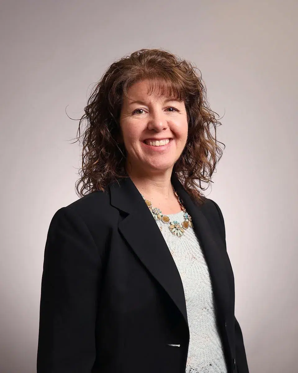 Woman with curly brown hair wearing a black blazer and light top, standing and smiling in front of a plain light background.