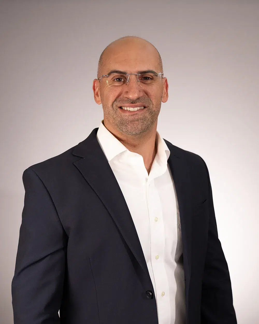 A man with glasses, wearing a dark suit jacket and white shirt, smiles at the camera against a plain light gray background.