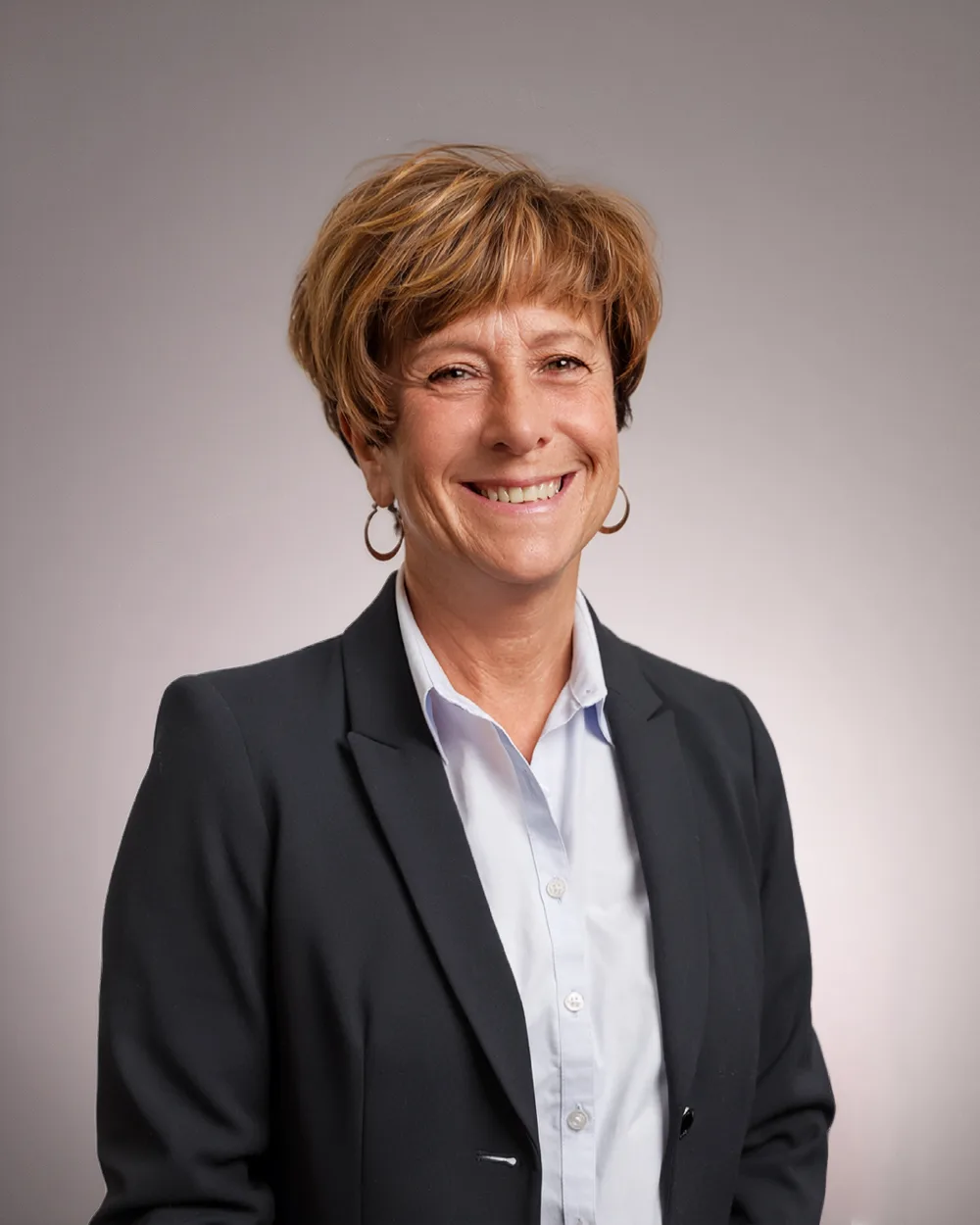 A woman with short brown hair, wearing a dark blazer and white shirt, smiles at the camera against a plain gray background.
