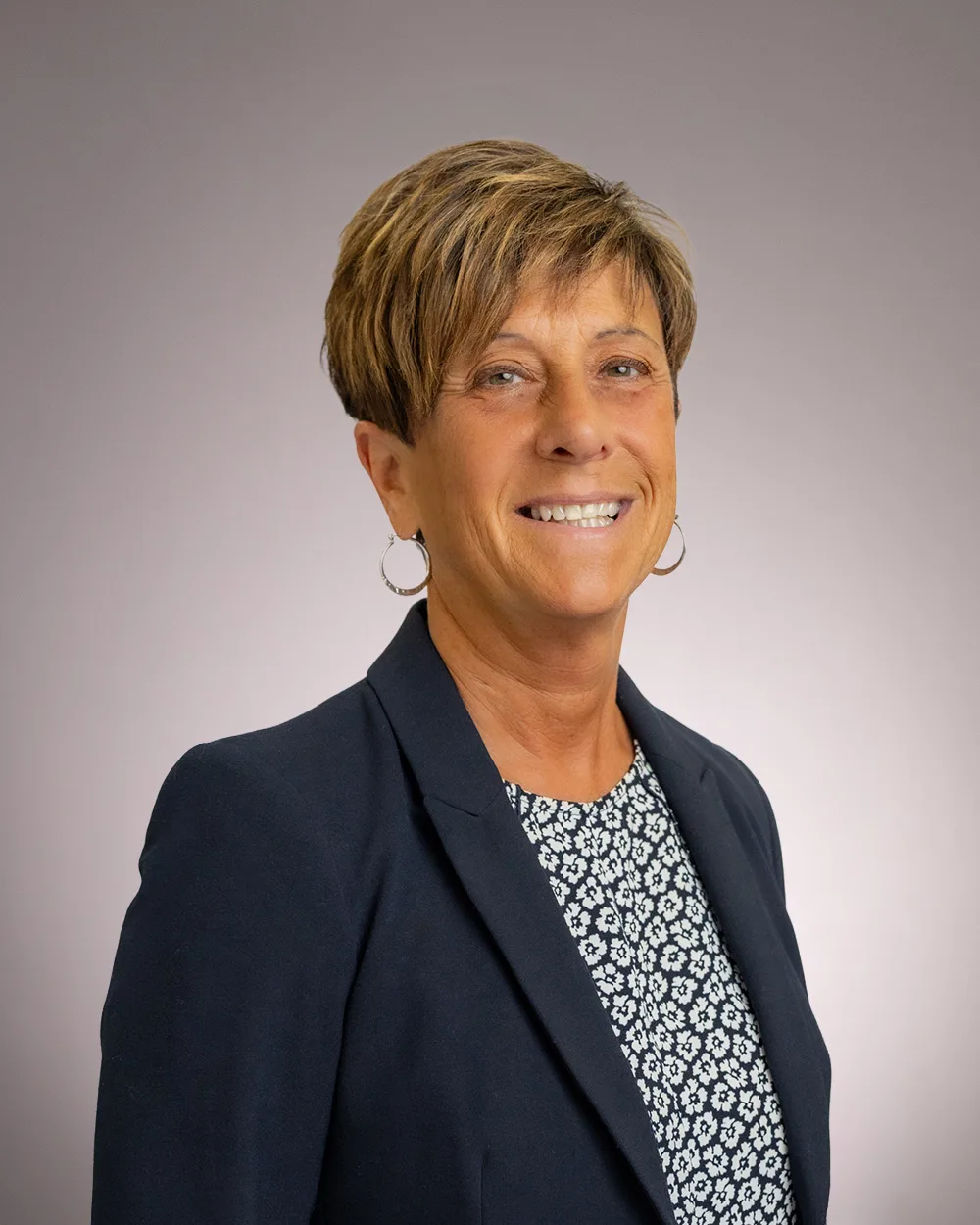 Smiling woman with short brown hair, wearing hoop earrings, a navy blazer, and a patterned shirt, standing in front of a plain, light gray background.