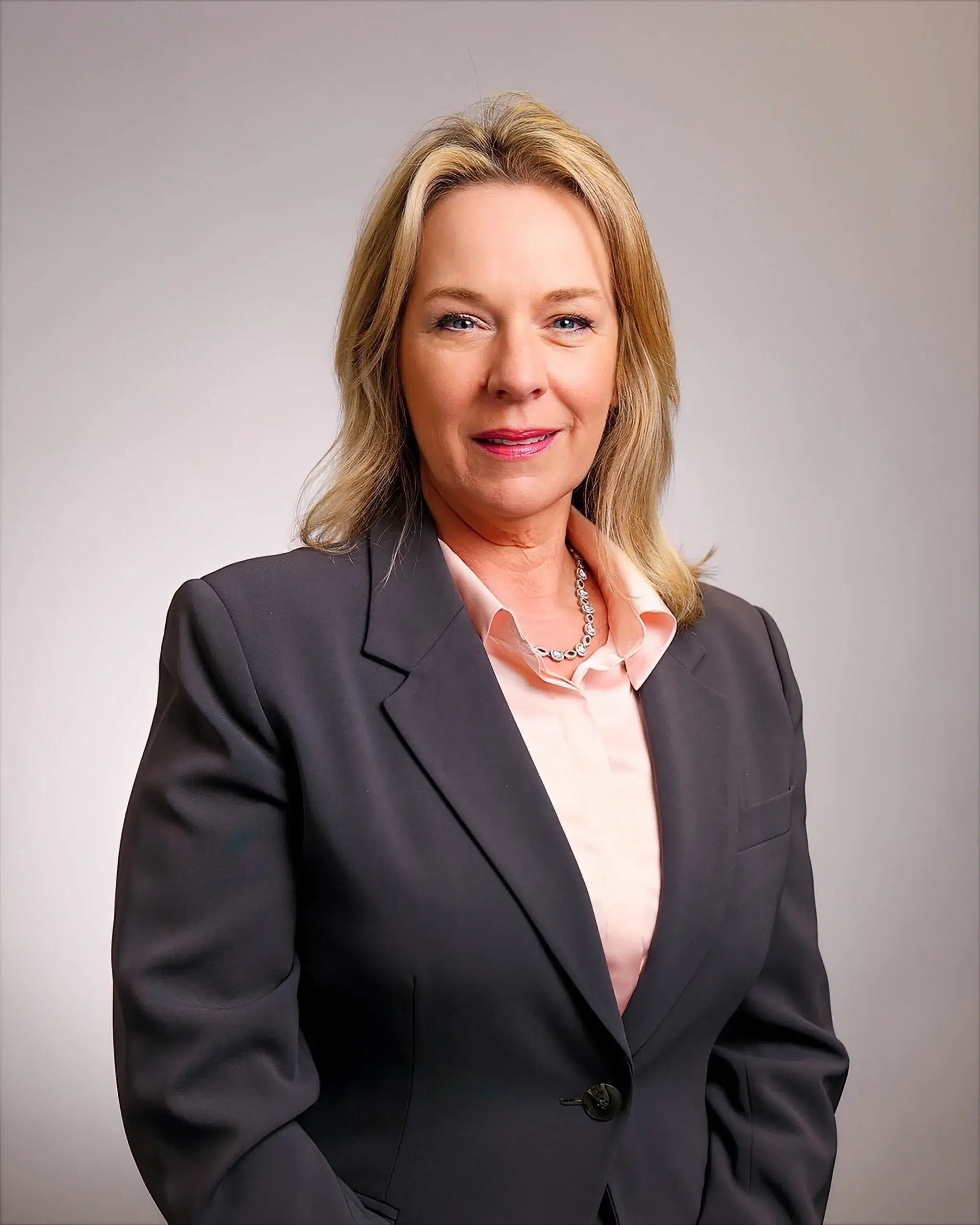 A woman with blonde hair wearing a dark blazer, light pink shirt, and necklace, poses in front of a plain light background, smiling slightly at the camera.