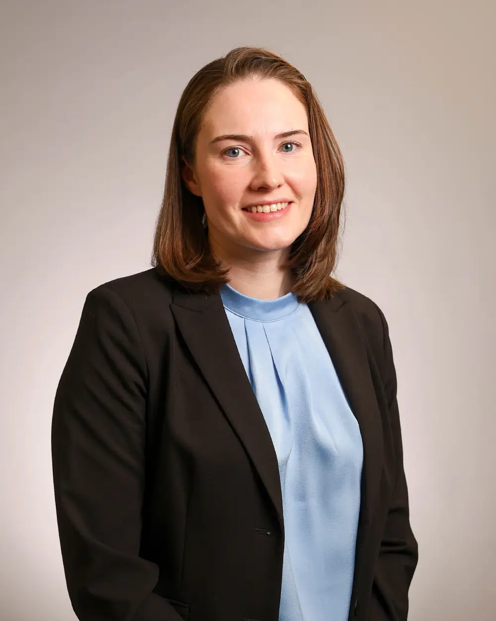 A woman with straight brown hair wearing a black blazer and a light blue blouse smiles at the camera against a plain light background.