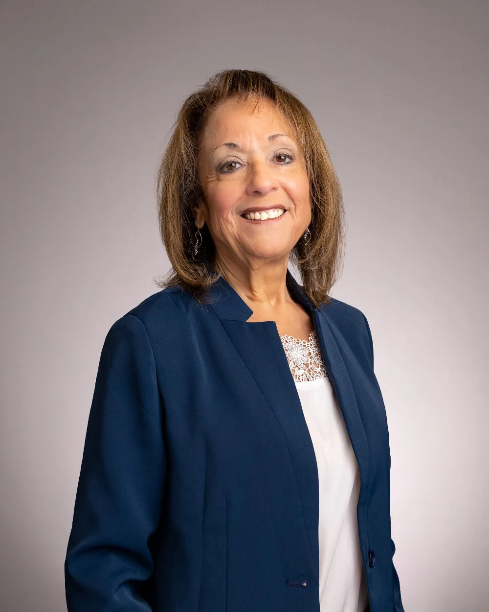 A woman with shoulder-length brown hair wearing a navy blazer and white blouse, smiling against a neutral gray background.