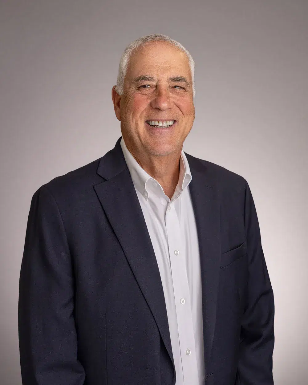 Smiling older man with short gray hair, wearing a dark suit jacket and white dress shirt, standing against a plain light gray background.