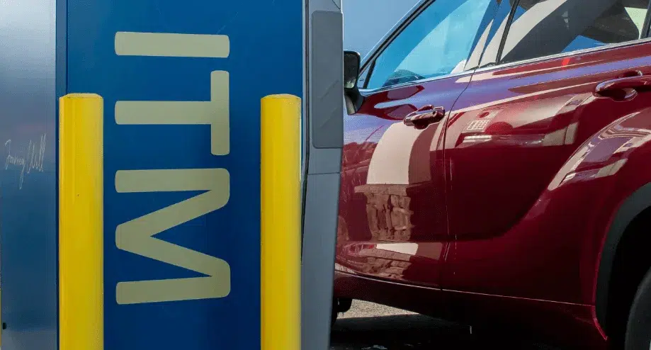 A red car parked next to a drive-thru ITM (Interactive Teller Machine) with yellow safety bollards in front of the machine.