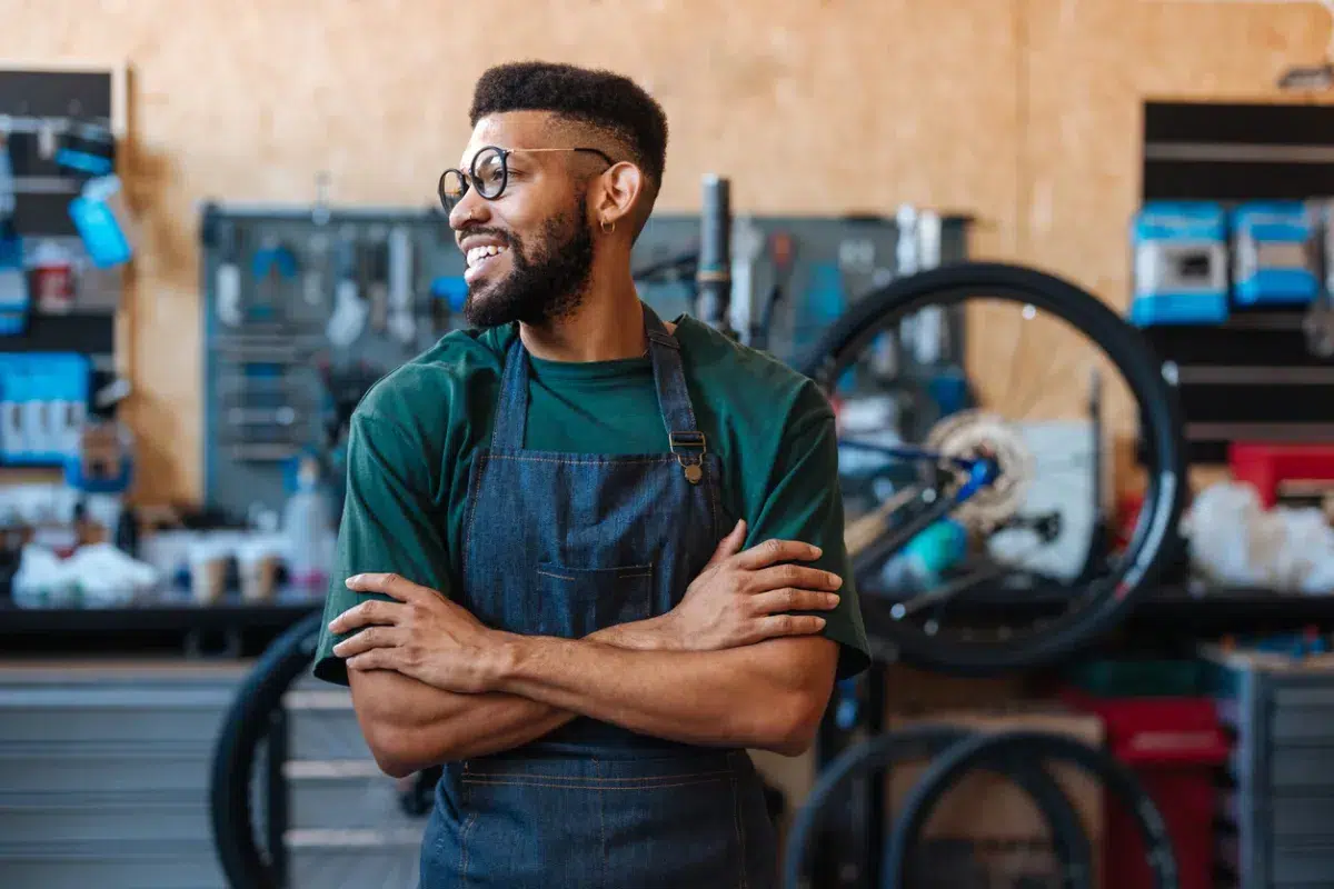 A smiling man in glasses and an apron stands with arms crossed in a bike repair shop, reflecting the hardworking spirit of commercial lending Massachusetts businesses.
