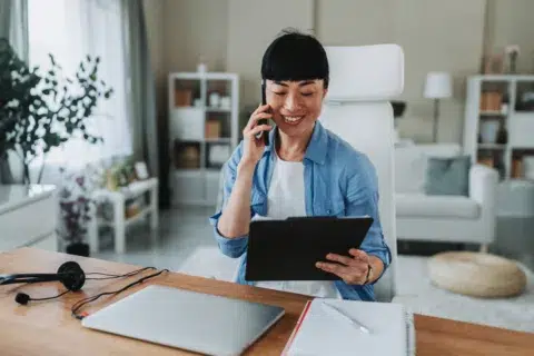 Woman sits at her home office desk, smiling while talking on the phone about commercial lending in Massachusetts, holding a tablet with a laptop and notebook nearby.