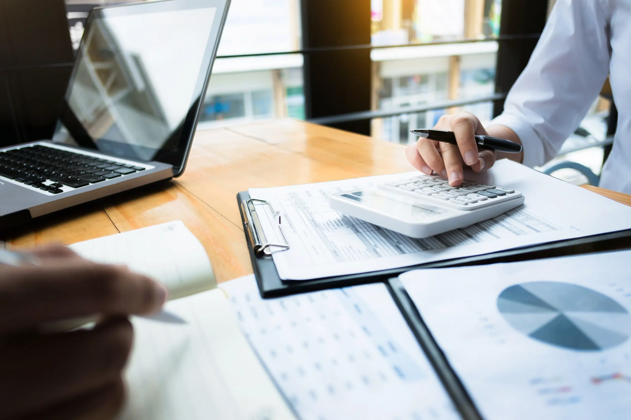Two people working at a desk with a laptop, calculator, documents, and charts, analyzing financial data and taking notes.