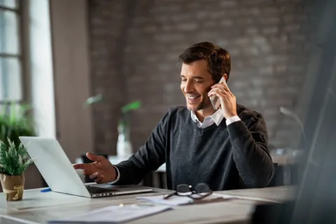 Smiling man in a sweater talks on a smartphone while working on a laptop at a desk in a bright office.