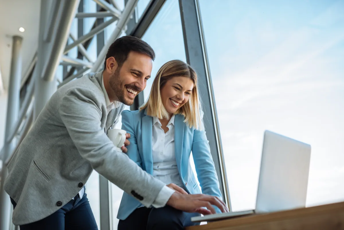 Two smiling people in business attire look at a laptop together by large windows in a modern office setting.