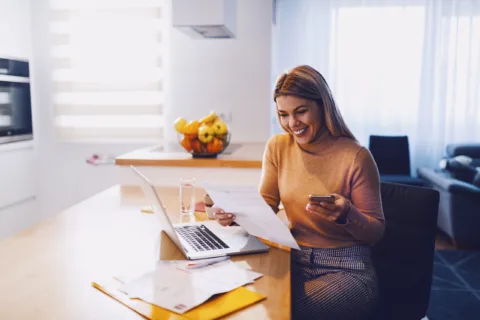 A woman sits at a kitchen table, smiling while holding a phone and a document. A laptop, papers, and a glass of water are on the table; fruit bowl in the background.