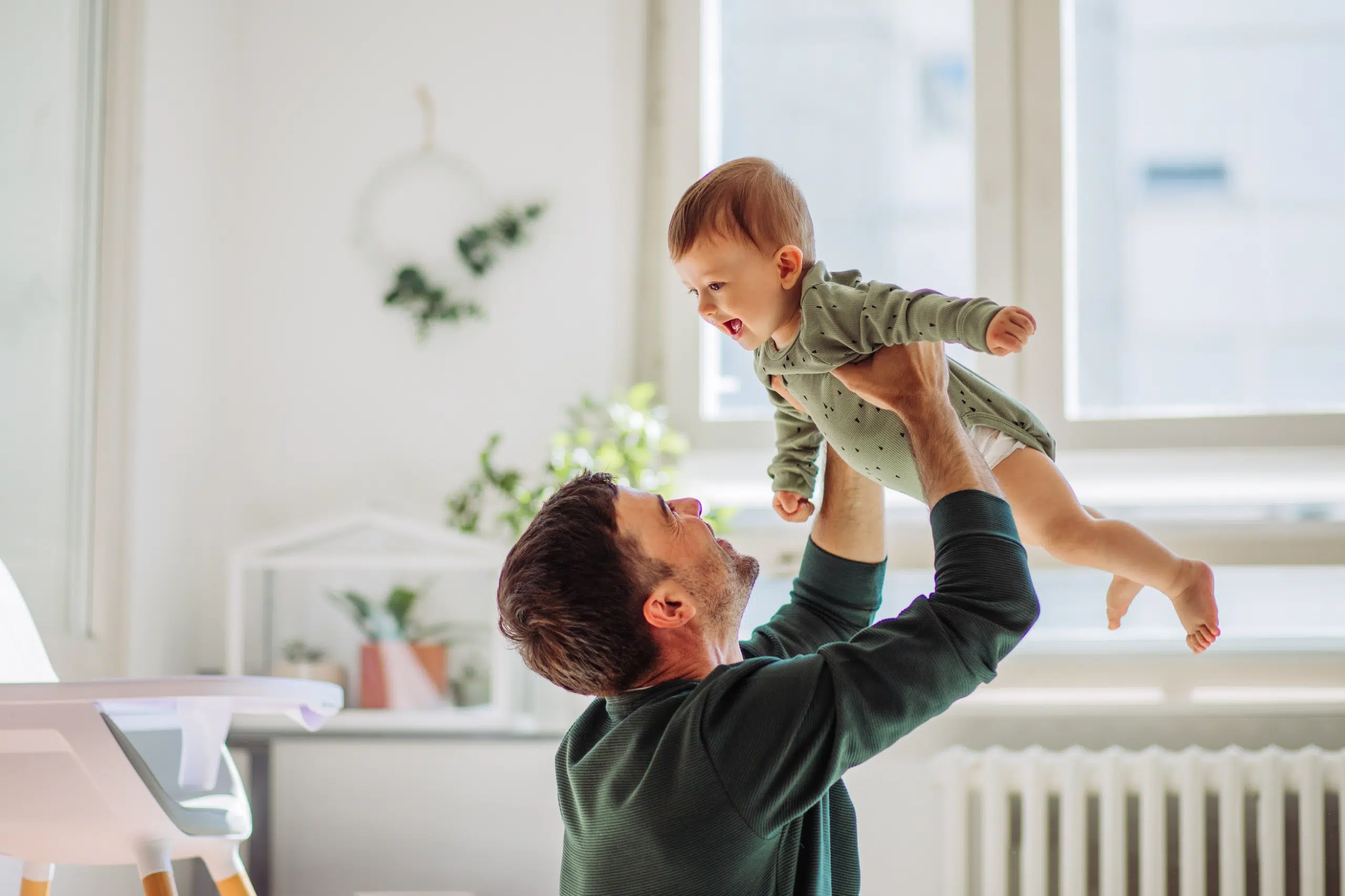 A man lifts a smiling baby in the air in a bright, cozy room with large windows and green plants in the background.